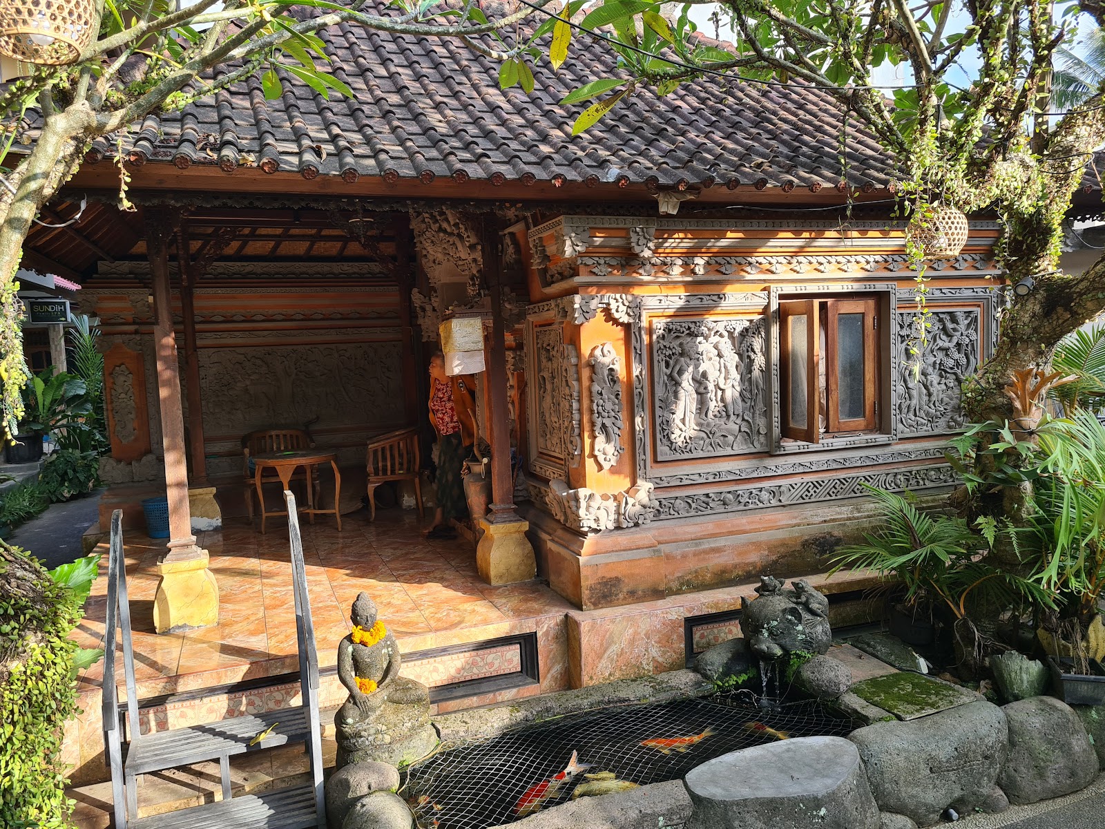 Traditional Balinese wooden structure with intricate carved details, dark tile roof, and courtyard featuring a decorative koi pond with stone statues. Lush tropical vegetation surrounds the property, creating an authentic and serene atmosphere.