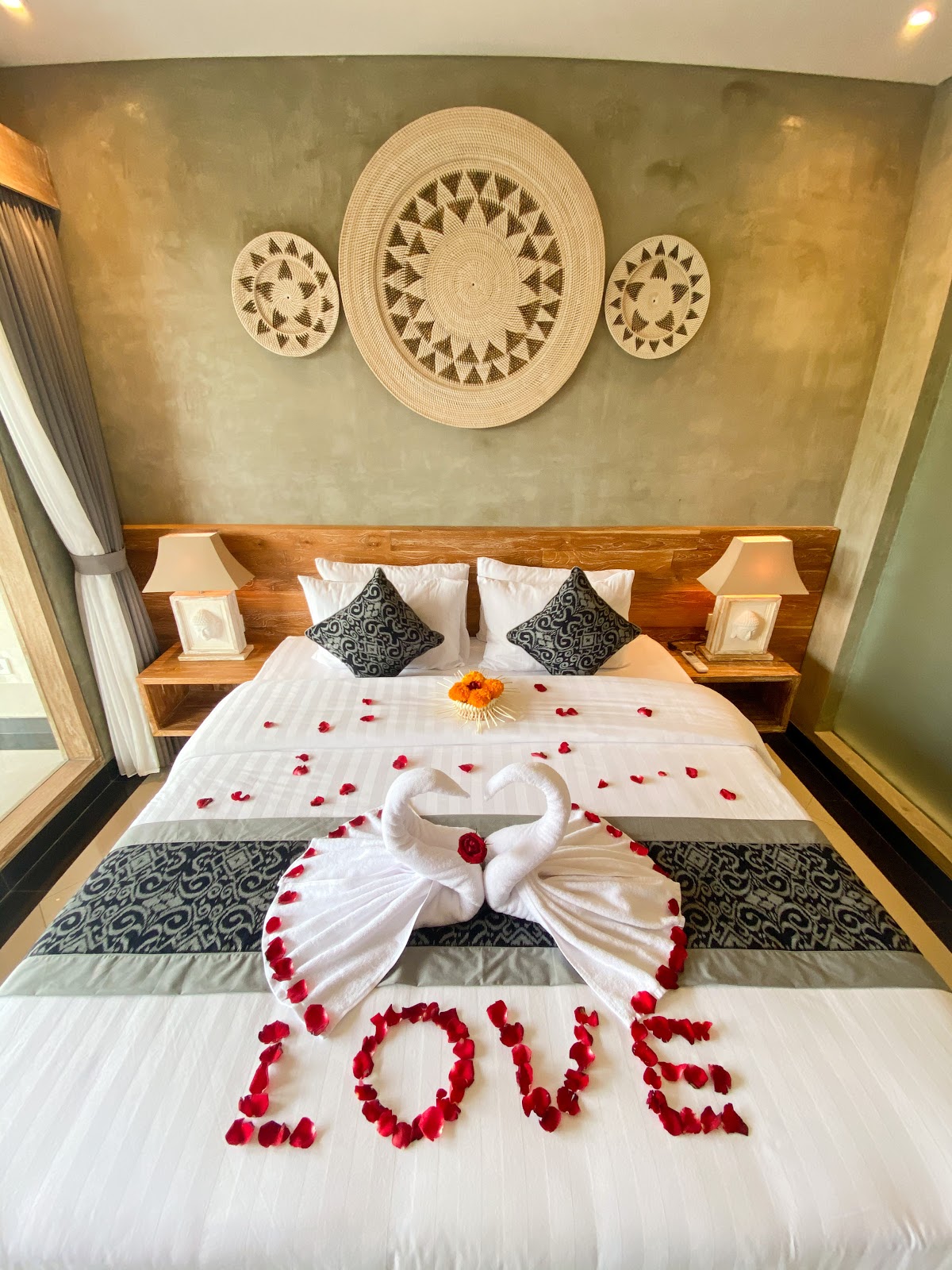 Romantic bedroom setup with heart-shaped towel arrangement, red rose petals spelling 'LOVE', decorative woven wall plates, wooden bed with patterned pillows, and ambient lighting creating an intimate atmosphere perfect for honeymoon or special occasions.