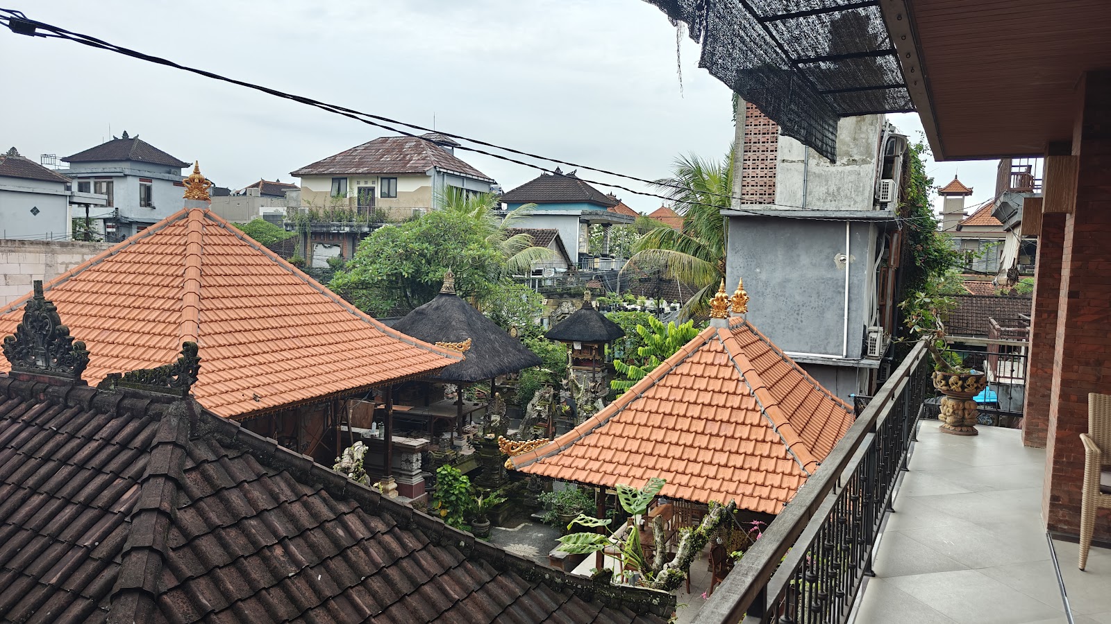 Aerial view of a Balinese homestay featuring traditional terracotta tile roofs with ornamental decorations, lush tropical vegetation, and multiple pavilion-style structures arranged around courtyards. The property shows characteristic Indonesian architecture with decorative roof finials and garden elements.